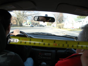 Photograph taken from the back of a car. In the driver's seat is a younger white person with gropped short hair and black glasses. In the passenger's seat is an older white person with white hair and glasses. The photographer holds a yellow tape deck down the middle horizontally.