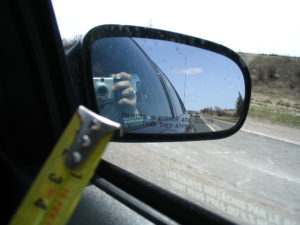 Photograph of a car's side-view mirror, taken from inside the car. Near the camera and slightly out of focus, a yellow tape measure can be seen. The mirror reflects a person holding up a camera to it. Written on the mirror are the words: 'Objects in mirror are closer than they appear'.