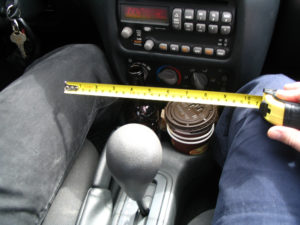 Photograph taken inside a car, showing the space between driver and passenger. In the middle is a gear stick and a cup holder with cups of coffee. The right leg of the driver is seen, wearing denim. The left leg of the passenger is seen, also wearing denim. The passenger is holding a yellow tape measure between them.