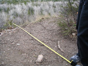 Photograph taken in the countryside, near the edge of a cliff. In the foreground, a person's legs and feet wearing gray trainers can be seen. From the person's feet to the edge, a yellow tape measure is extended.