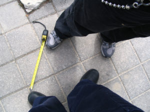 Photograph looking down, showing the legs and feet of two people standing on a tiled floor outside, one above and one below. Both people are wearing denim trousers. The person at the bottom is wearing black boots, the person at the top is wearing white and gray trainers. Between them to the left side is an open, yellow tape measure.
