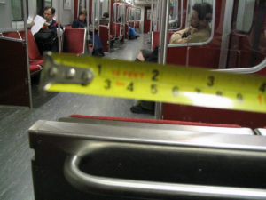 Photograph taken inside a subway carriage. The carriage is about a third full, with several people sitting down. A yellow tape measure is held horizontally, close to the camera, measuring the distance to the nearest passenger. The seats are a mixture of bright red cloth and stainless steel.