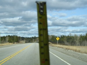Photograph of a highway in the countryside, near a forest. The upper half of the picture shows a cloudy sky. A yellow tape measure is held vertically in the middle, close to the camera and slightly darkened and out of focus.