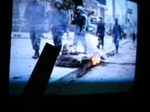 The photograph shows a TV screen, which is showing footage of soldiers during a war. An object is on fire in the foreground of the footage. A tape measure is held diagonally towards the middle, obscured and in silhouette.