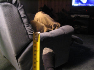 The photo depicts a person curled up in a couch and watching TV, seen from the side. Only the person's legs are visible. In the foreground, the photographer's right foot is seen wearing a sock. A yellow tape measure is held up in the middle, slightly out of focus.