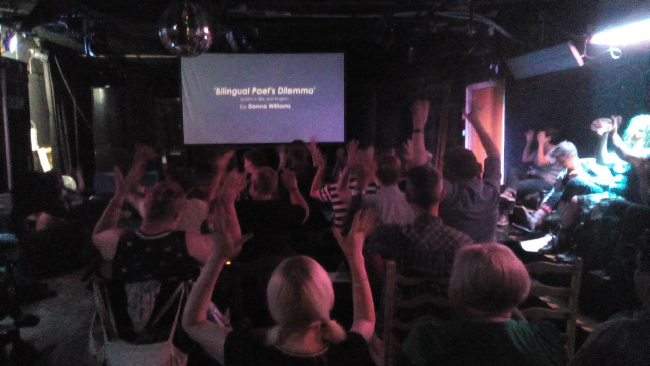 Photo image of an audience from behind, facing a screen with the title card 'Bilingual Poet's Dilemma by DL Williams'. Everyone in the audience has their hands raised, the BSL sign for 'clapping'.