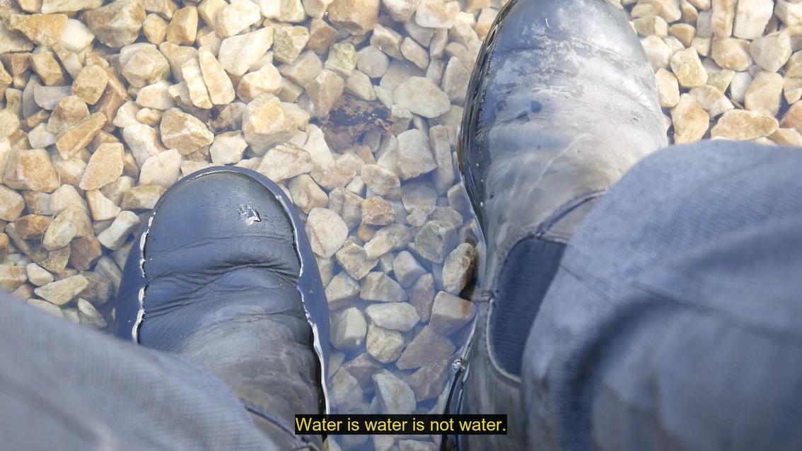 The image is from the point of view of someone sitting outside and looking down at their feet. The view is of a deep and reflective clear rain puddle on ground made of large, orangey pebbles. The person is wearing grey trousers and has worn, black boots half-covered in water. A caption reads: Water is water is not water.