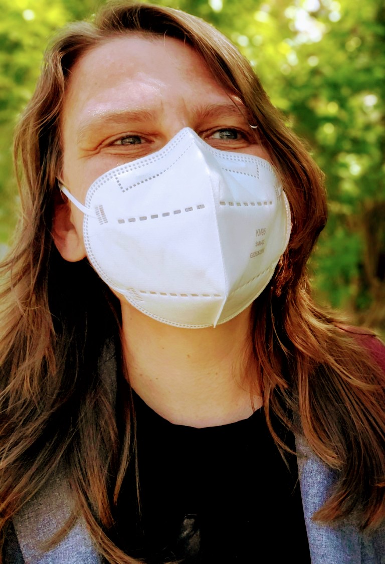 A close-up of San outside wearing a white N95 mask, black t-shirt and grey jacket. San is white with long reddish-brown hair, smiling with crinkly blue eyes at someone off-camera. Green trees with sun-dappled leaves blur behind them.