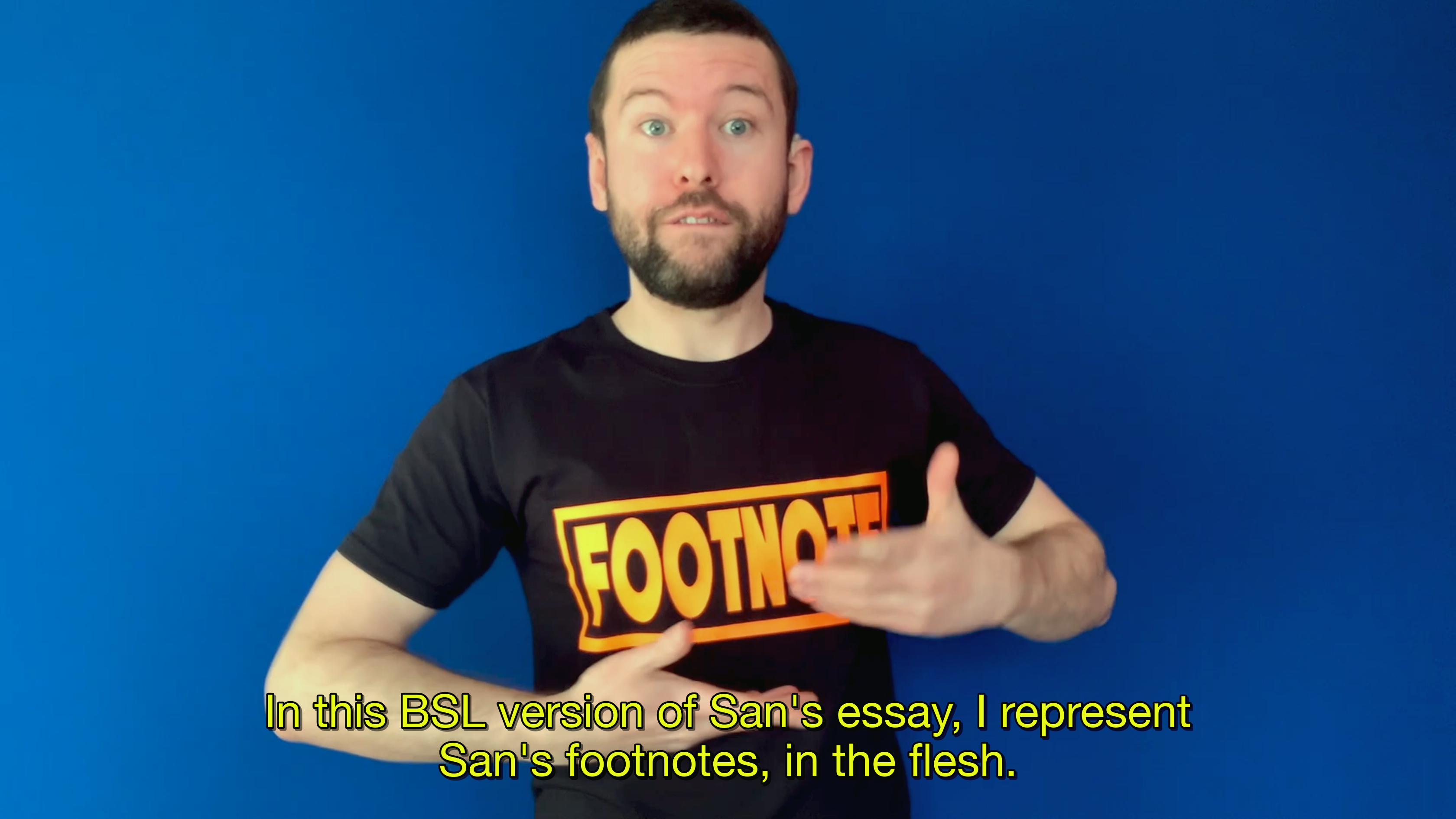 A video still from San Alland's essay, of a white deaf person who stands in front of a bright blue wall. He's wearing a black t-shirt that says "Footnote" in bright orange letters. Yellow captions say: 'In this BSL version of San's essay, I represent San's footnotes, in the flesh.'