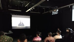 A group of people, some wheelchair-users, and all wearing respirators, watch a film.